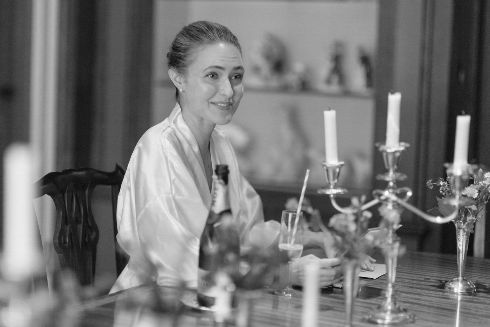 A woman with a pearl earring and hair tied back, sitting at a dining table with candlesticks, a bottle, and a glass, smiling.