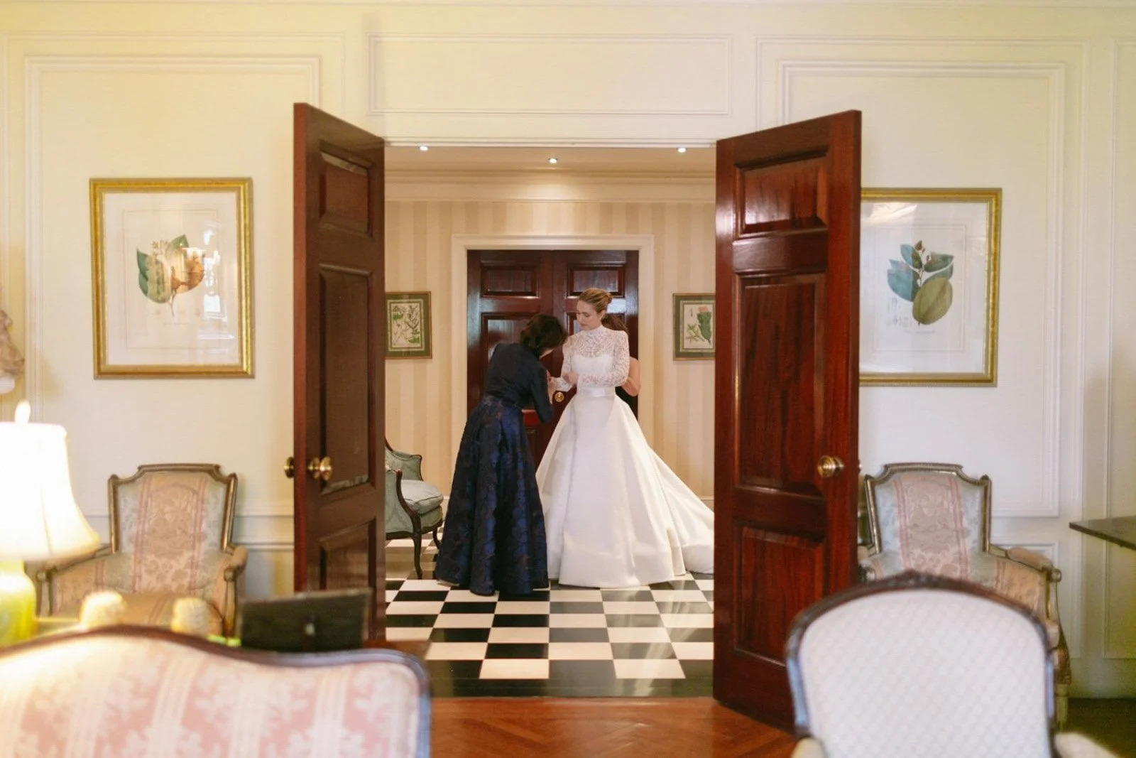 A woman in a white wedding gown standing in a room as another woman adjusts her dress.