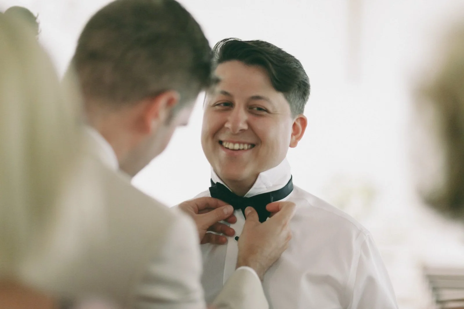 A man with dark hair wearing a white dress shirt smiles as another person is adjusting his black bow tie.