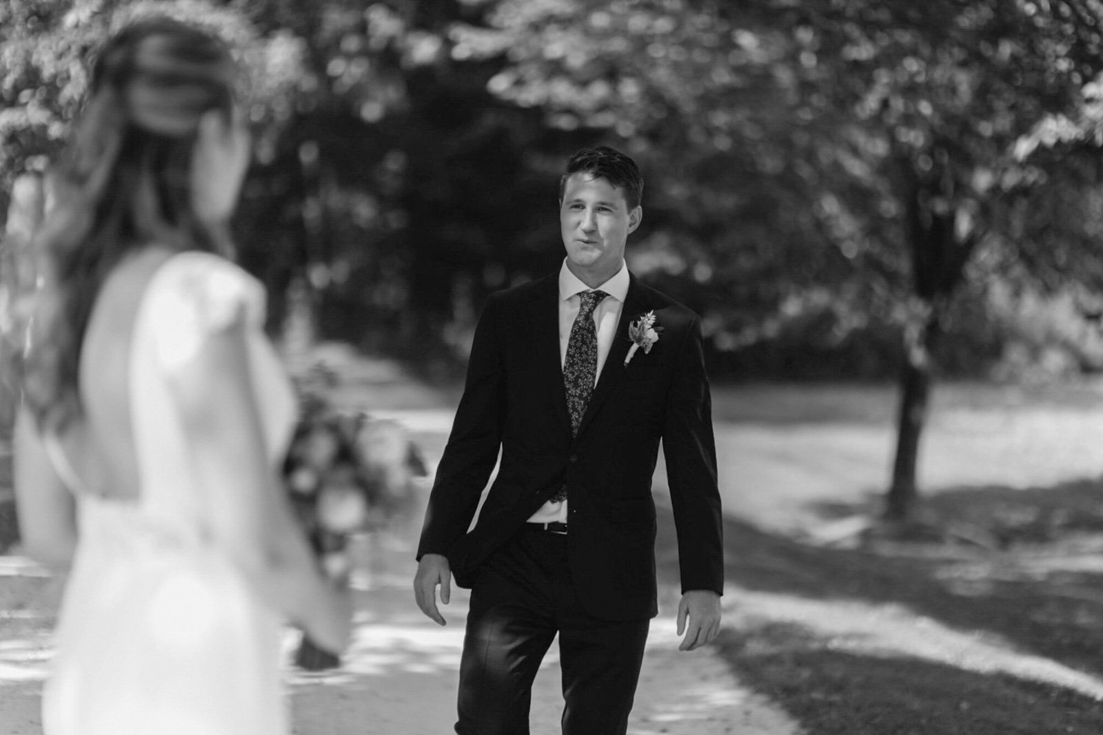 A man in a suit standing outdoors on a sunny day, looking at a woman dressed in white who is slightly out of focus in the foreground at Court Hill in The Berkshires.