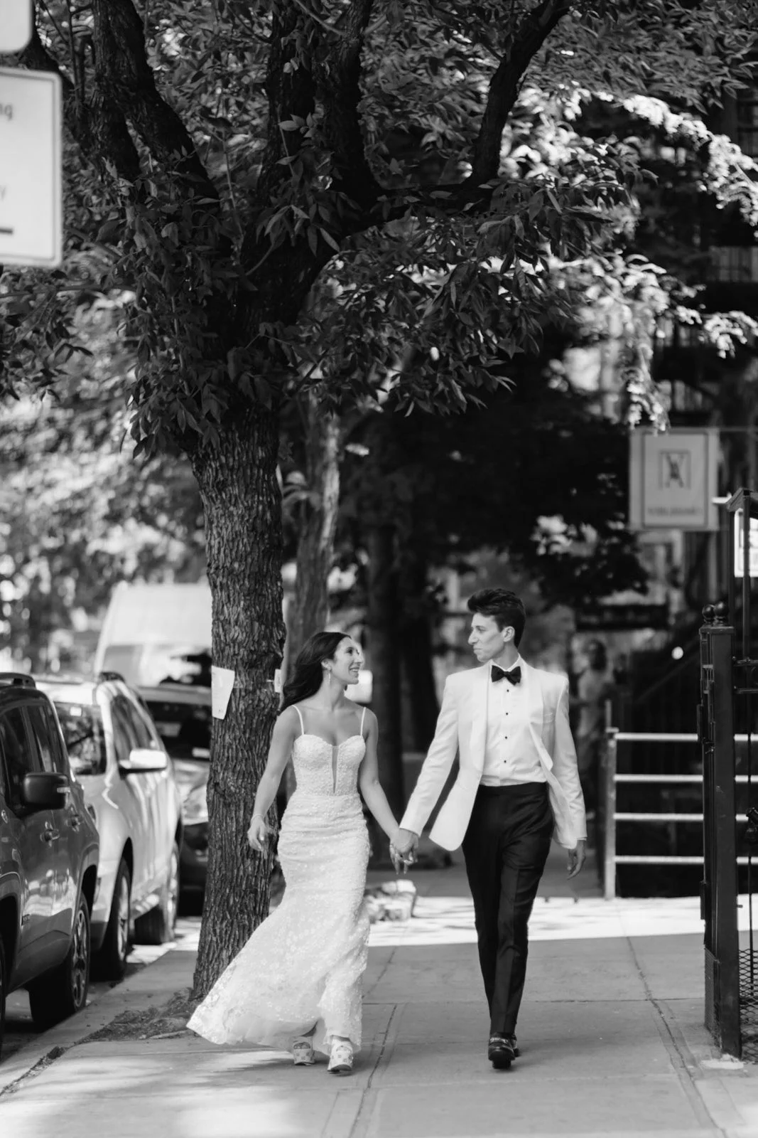 A bride and groom walking hand in hand on a city sidewalk, smiling and looking at each other, with parked cars and trees lining the street.