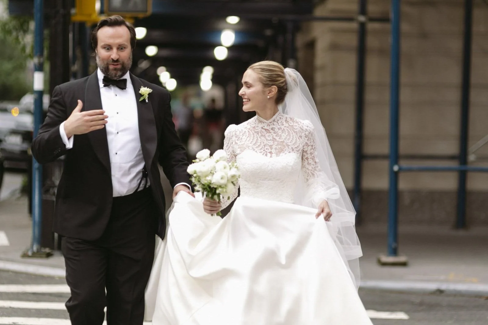 A bride and groom in wedding attire walking on a city street in the evening, smiling and holding flowers, engaged in conversation.