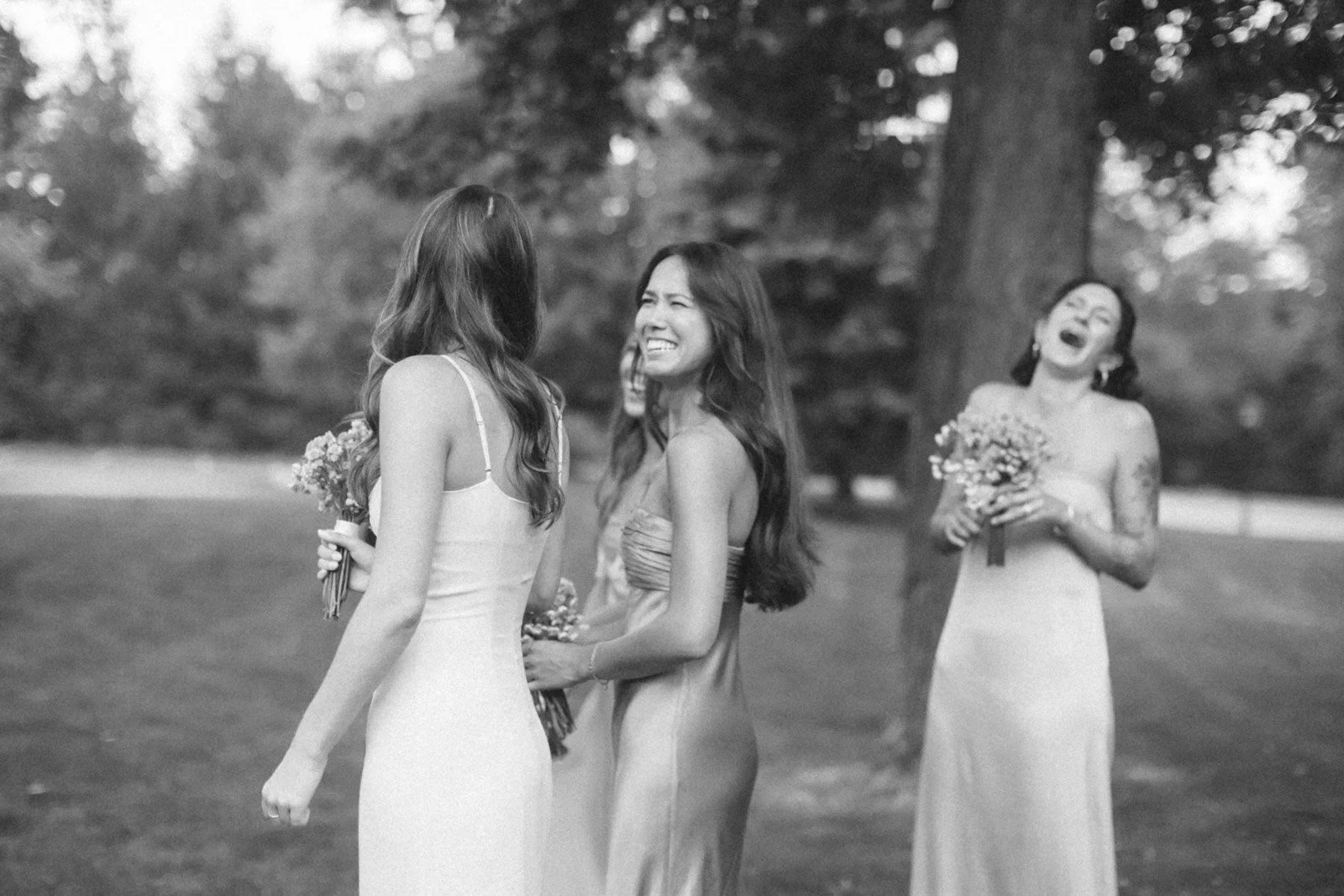 Two women in wedding dresses sharing a joyful moment, with another woman in a wedding dress in the background holding a bouquet, outdoors in a park with trees.