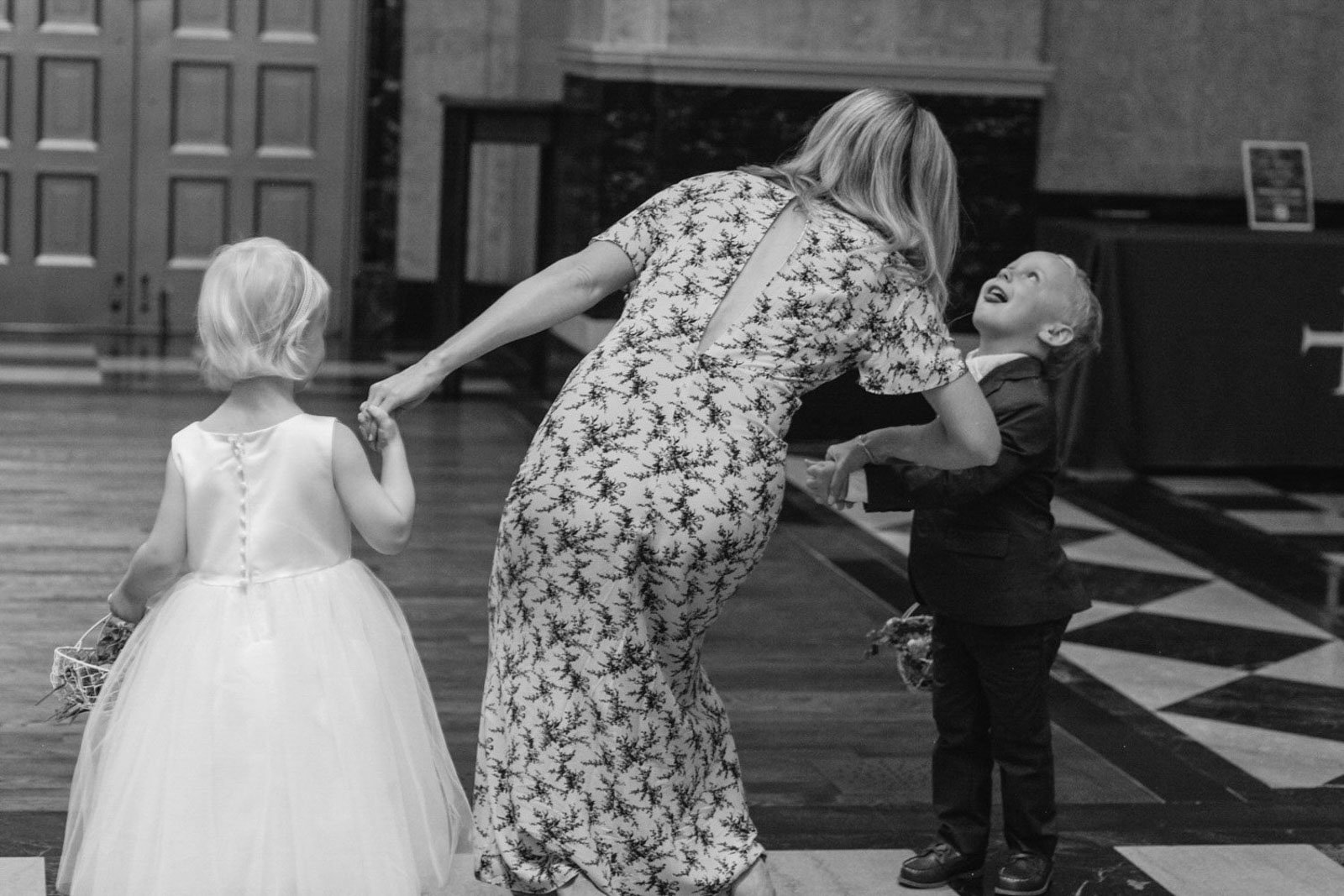 A woman in a patterned dress holding hands with a young girl in a white dress, while a young boy in a dark suit laughs and leans back towards her.