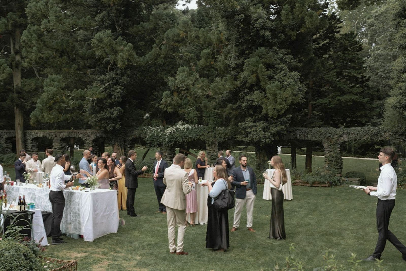 People gathered outdoors for a social event, standing in small groups, chatting, and drinking drinks, with a long white table with beverages on it, against a backdrop of lush green trees and a stone wall.