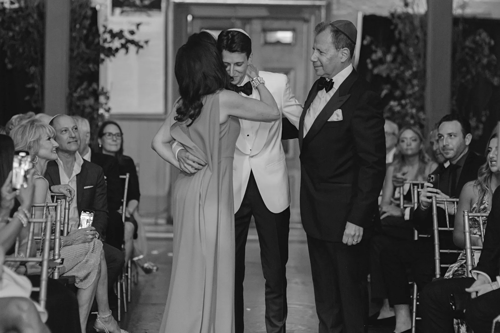 A wedding ceremony with a bride and groom standing together, surrounded by seated guests under indoor lighting. The bride is wearing a sleeveless gown and the groom is in a tuxedo with a bow tie. A woman in a dress appears to be emotional, possibly c