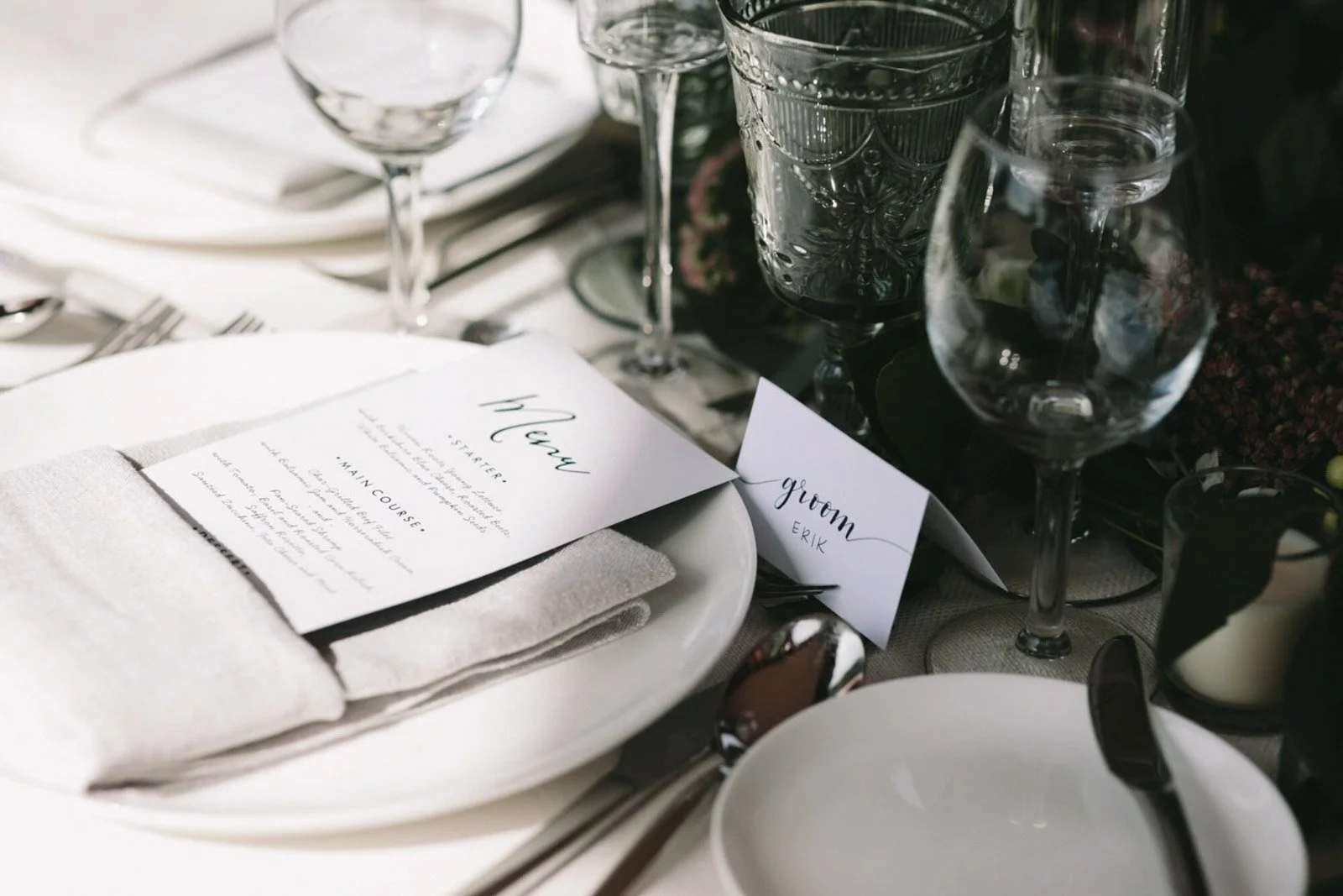 Elegant wedding table setting with white plates, cloth napkins, multiple glassware, and place cards labeled "Groom Erik" and "Bridgroom".