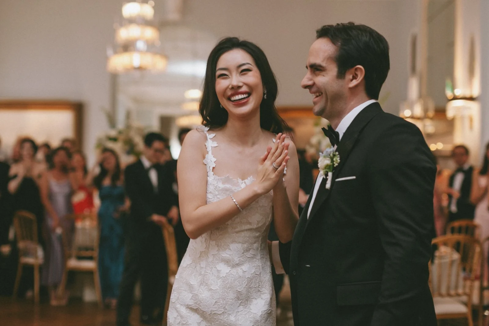 A bride in a white lace wedding dress smiling and holding hands with a groom in a black tuxedo during their wedding reception.