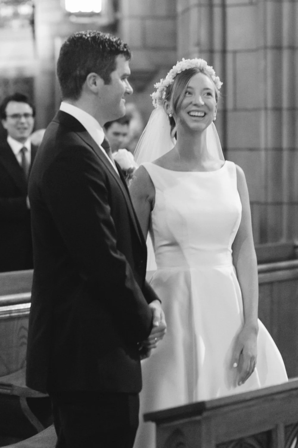A bride and groom standing during a wedding ceremony inside a church, with wedding guests visible in the background.