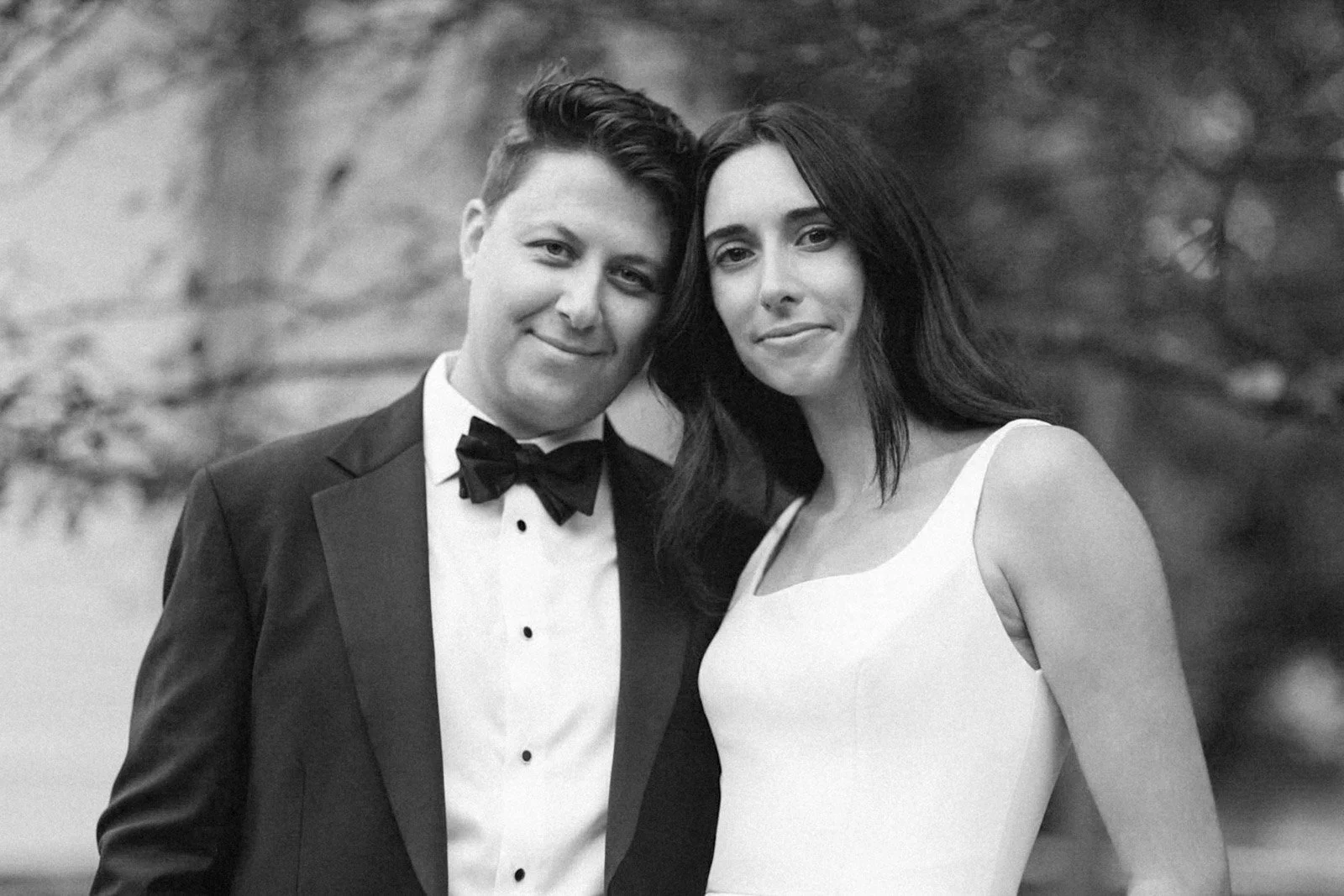 A black and white photo of a man in a tuxedo with a bow tie and a woman in a sleeveless dress, both smiling and standing close together outdoors.