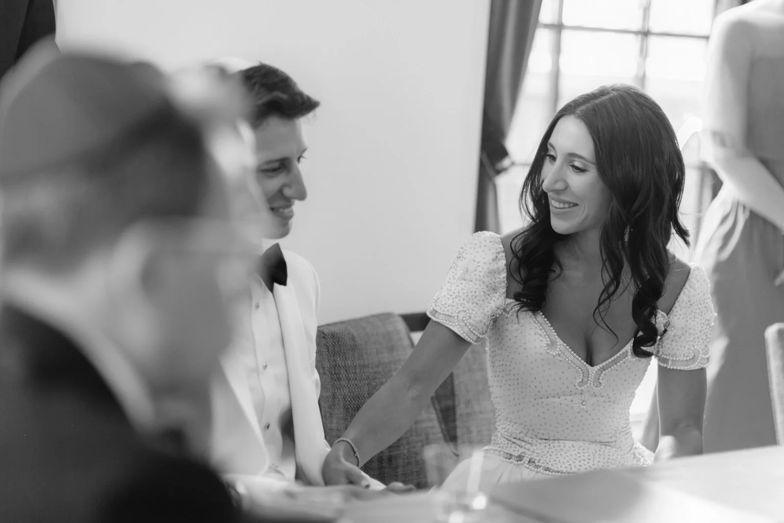 Black and white photo of a woman and two men sitting at a table, smiling and talking, with a window in the background.