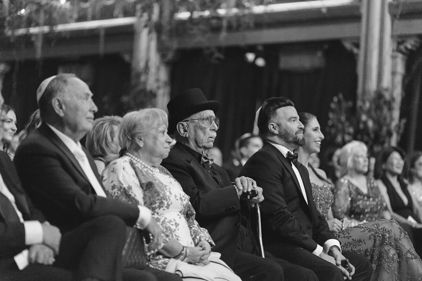Black and white photo of a formal event with several people seated in the audience, including an elderly man with a cane, a woman in a floral dress, and others in tuxedos and elegant dresses, watching attentively.