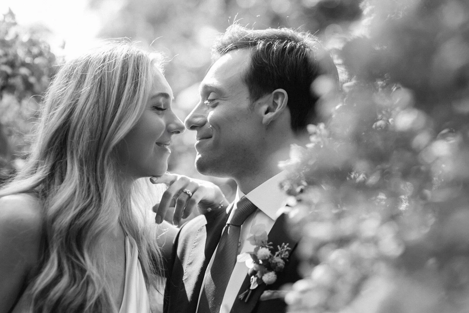 Black and white photo of a happy couple, a woman and a man, close to each other with their faces touching, smiling with eyes closed, outdoors with blurred foliage around them.