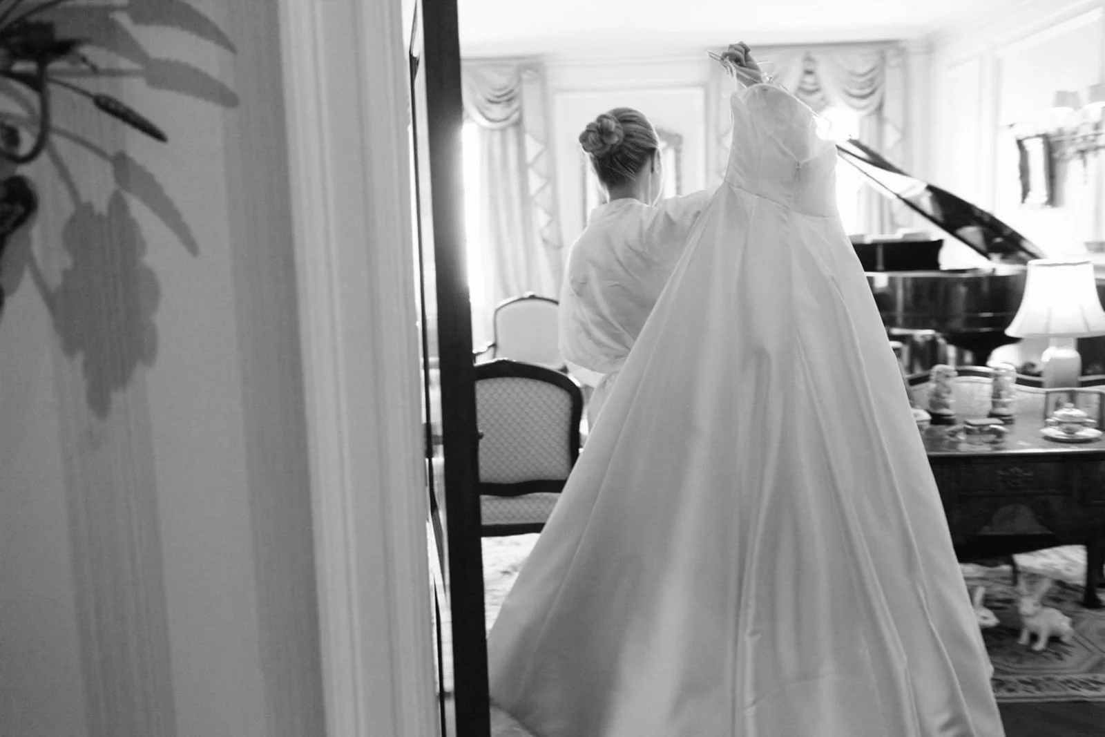 A woman with her hair in a bun preparing a wedding dress in a luxurious room with vintage furniture and patterned curtains.