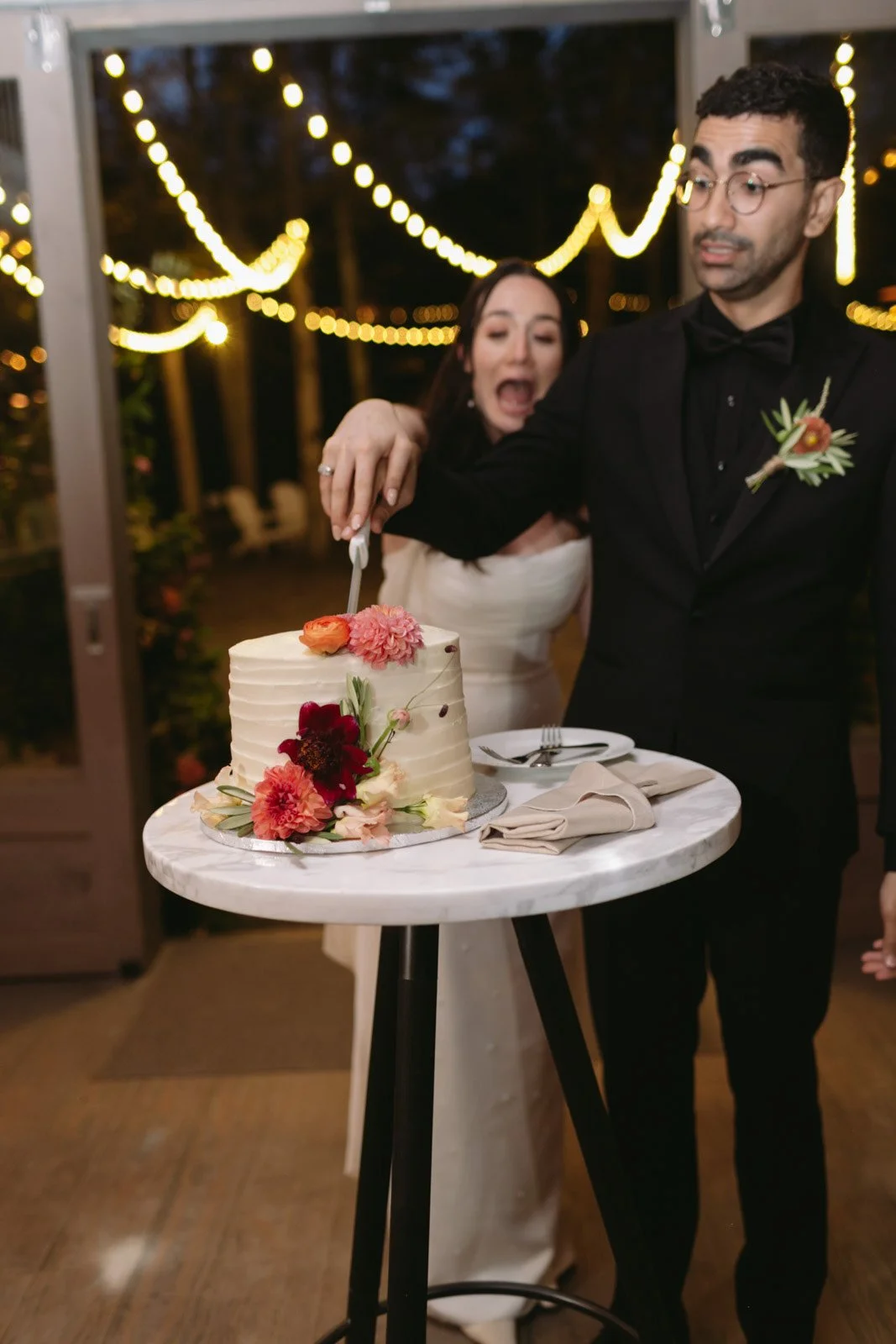 A newlywed couple is cutting their wedding cake decorated with flowers. The bride is in white, and the groom in a black suit with glasses, at nighttime with string lights in the background.