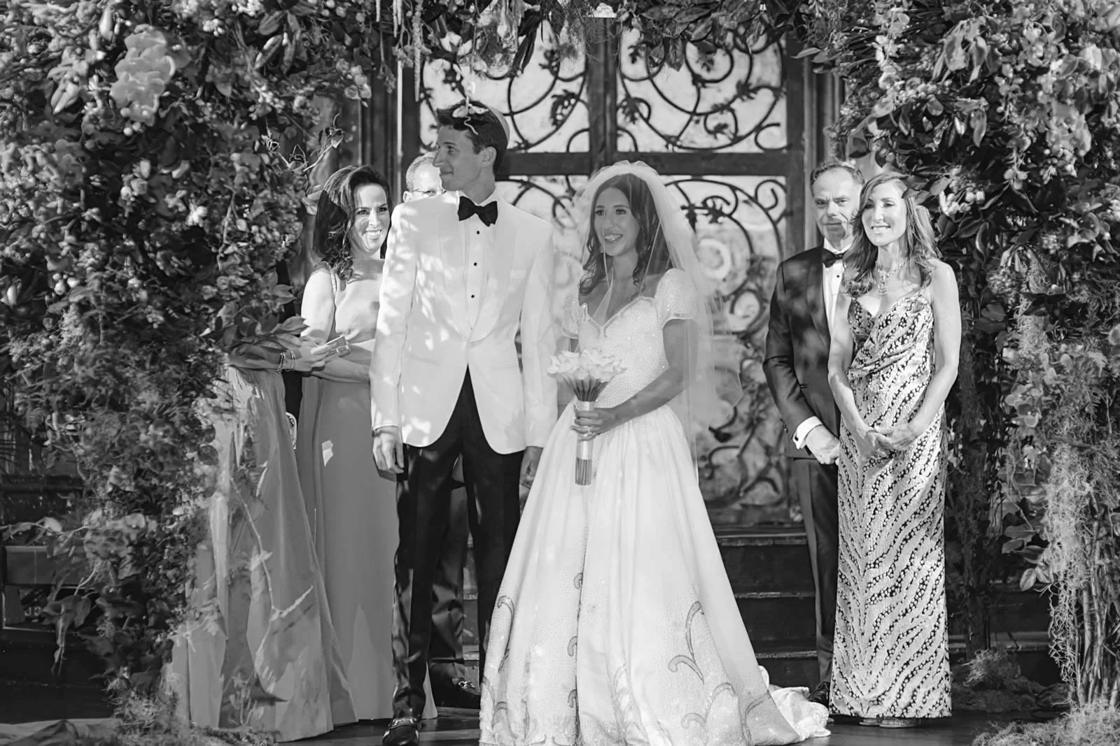 A black and white photo of a wedding ceremony with a bride in a wedding gown and veil holding a bouquet, and a groom in a tuxedo standing next to her. They are surrounded by six people, with a decorative iron gate and floral arrangements in the backg