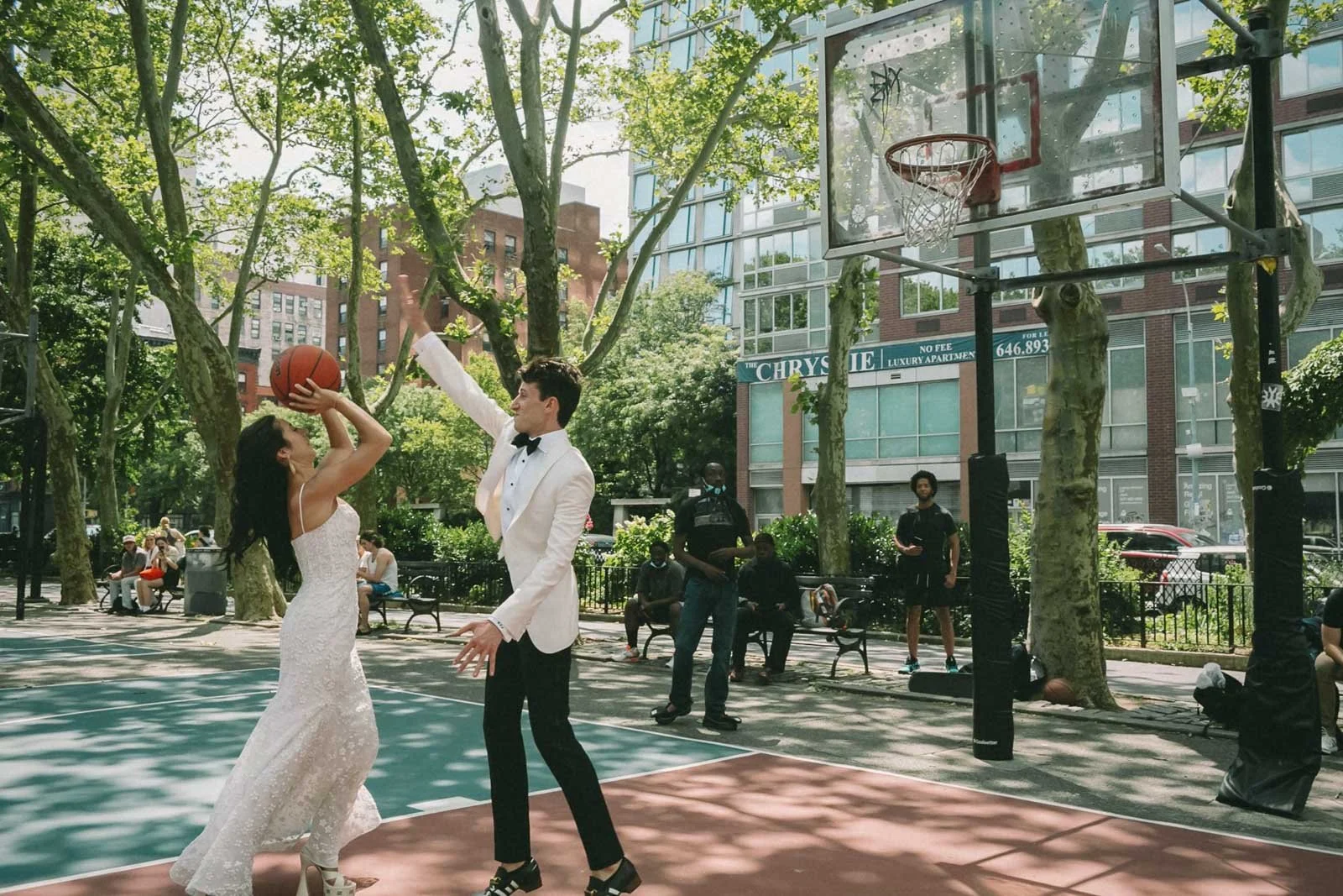 A couple dressed in formal attire playing basketball on an outdoor court in a park with trees and people in the background.