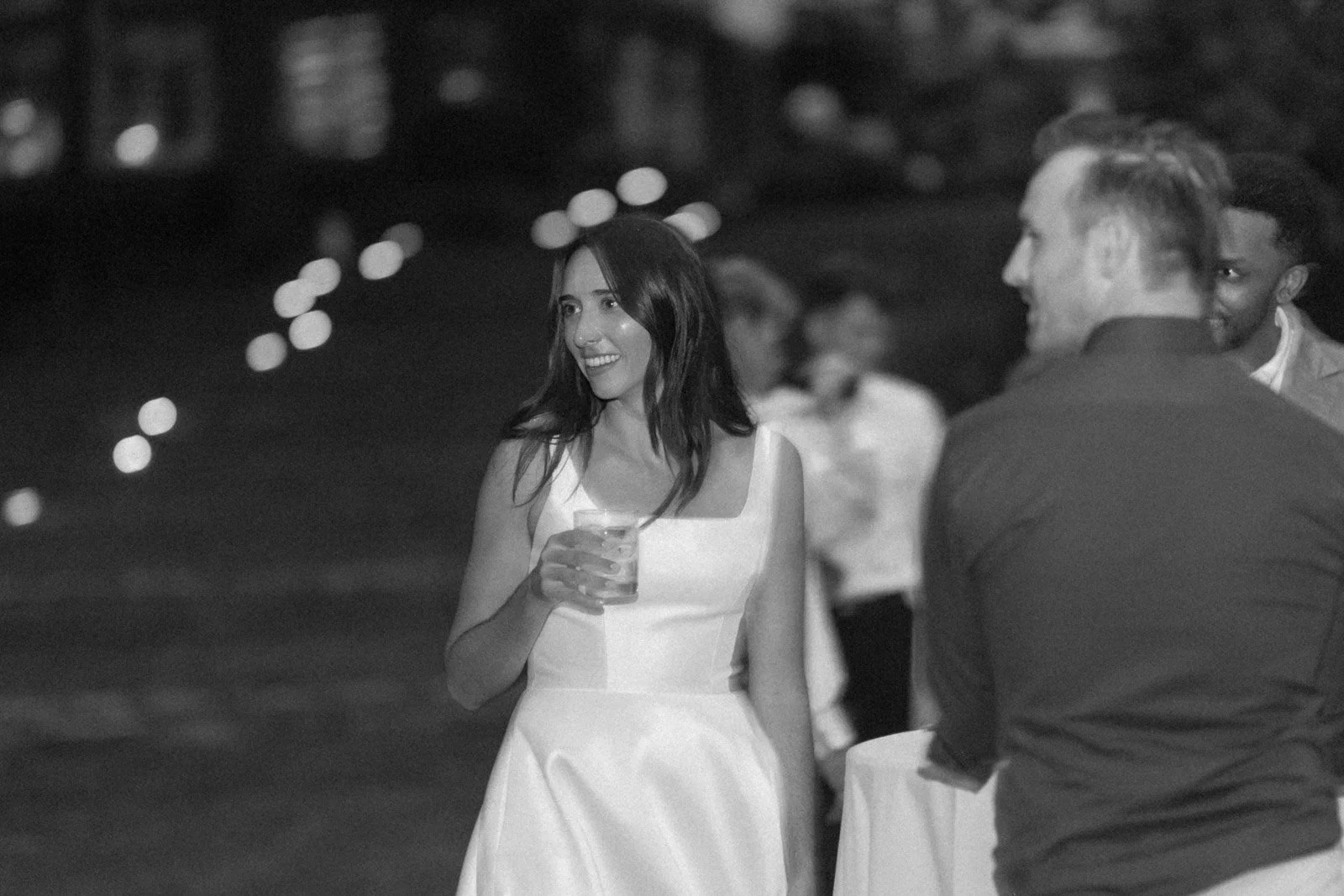 A woman in a sleeveless white dress smiling and holding a drink, standing at an outdoor event at night, with blurred lights and people in the background.