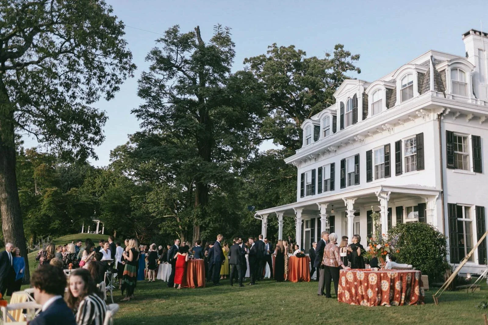 People gathered on the lawn outside a large white historic house for a social event or party during daytime, with some standing and others seated. Several high tables are set up with orange tablecloths, and there are trees and greenery in the backgro