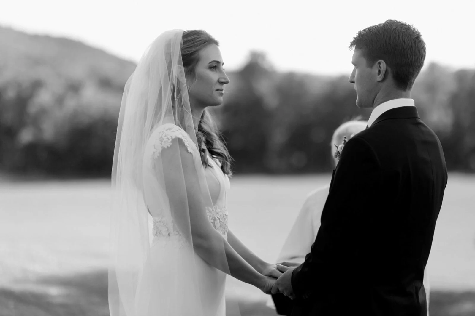A bride and groom holding hands during an outdoor wedding ceremony, facing each other, with a blurred natural landscape in the background in black and white.