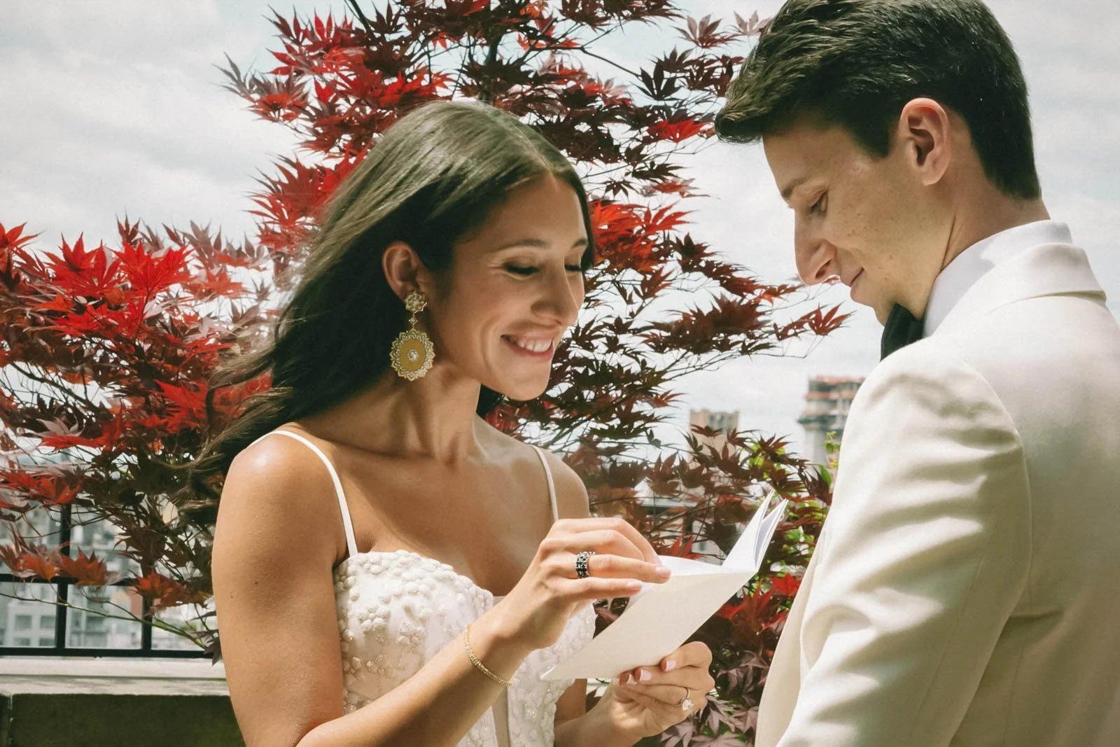 A woman in a white dress and big earrings is smiling while reading vows to a man in a white tuxedo, outdoors with red leaves in background.