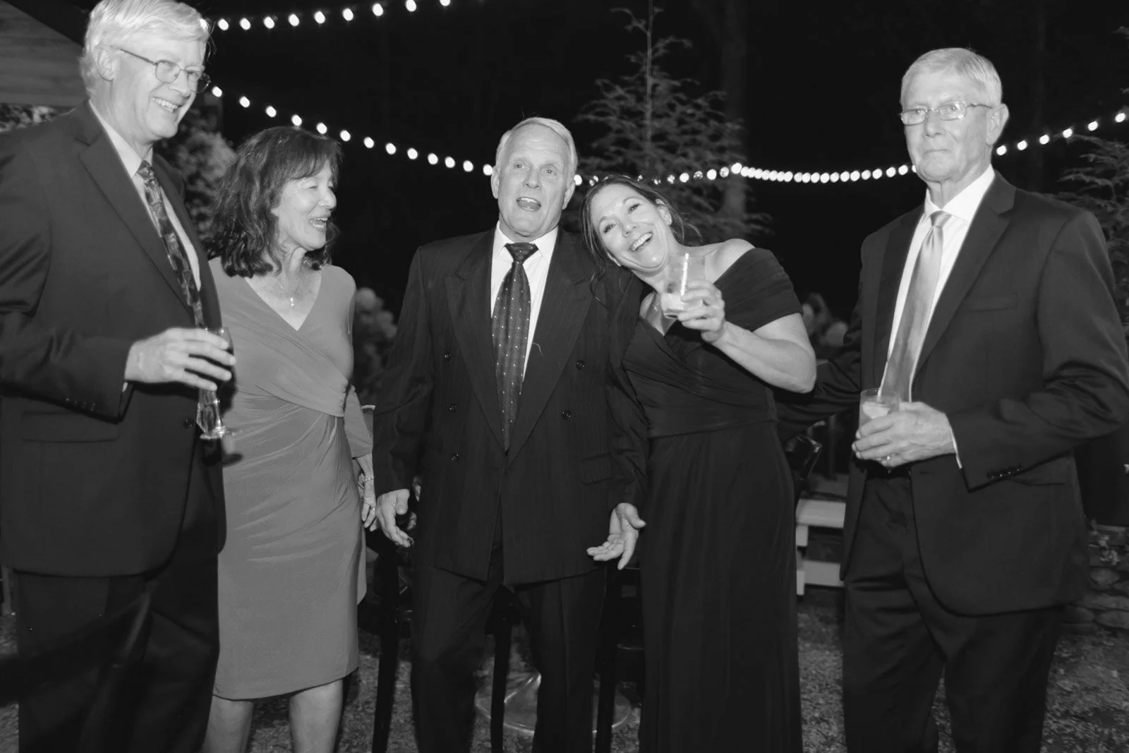 Group of five adults in formal attire at night, celebrating at an outdoor event with string lights overhead. They are holding drinks and smiling.