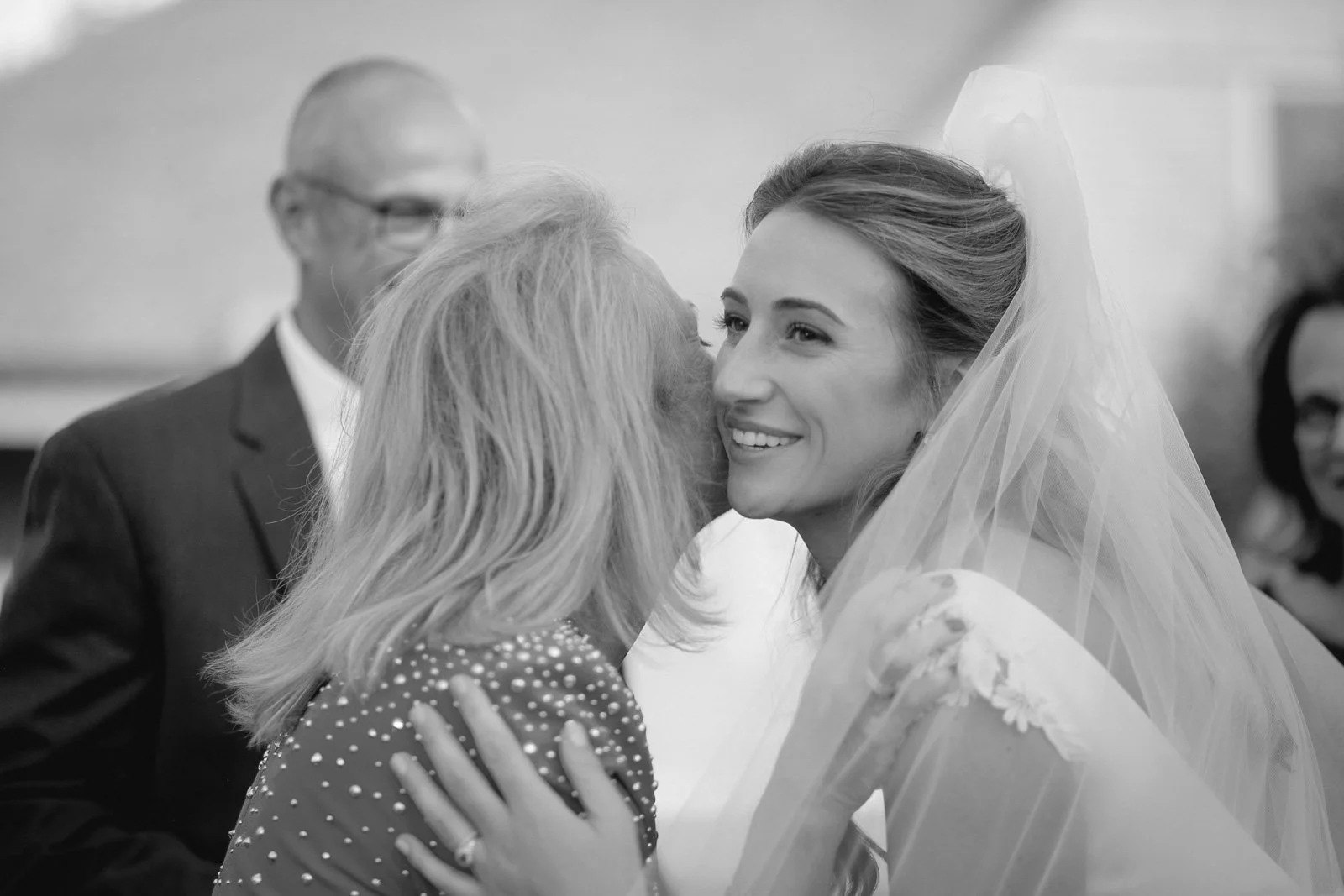 A bride smiling as she is greeted by an older woman during her wedding.