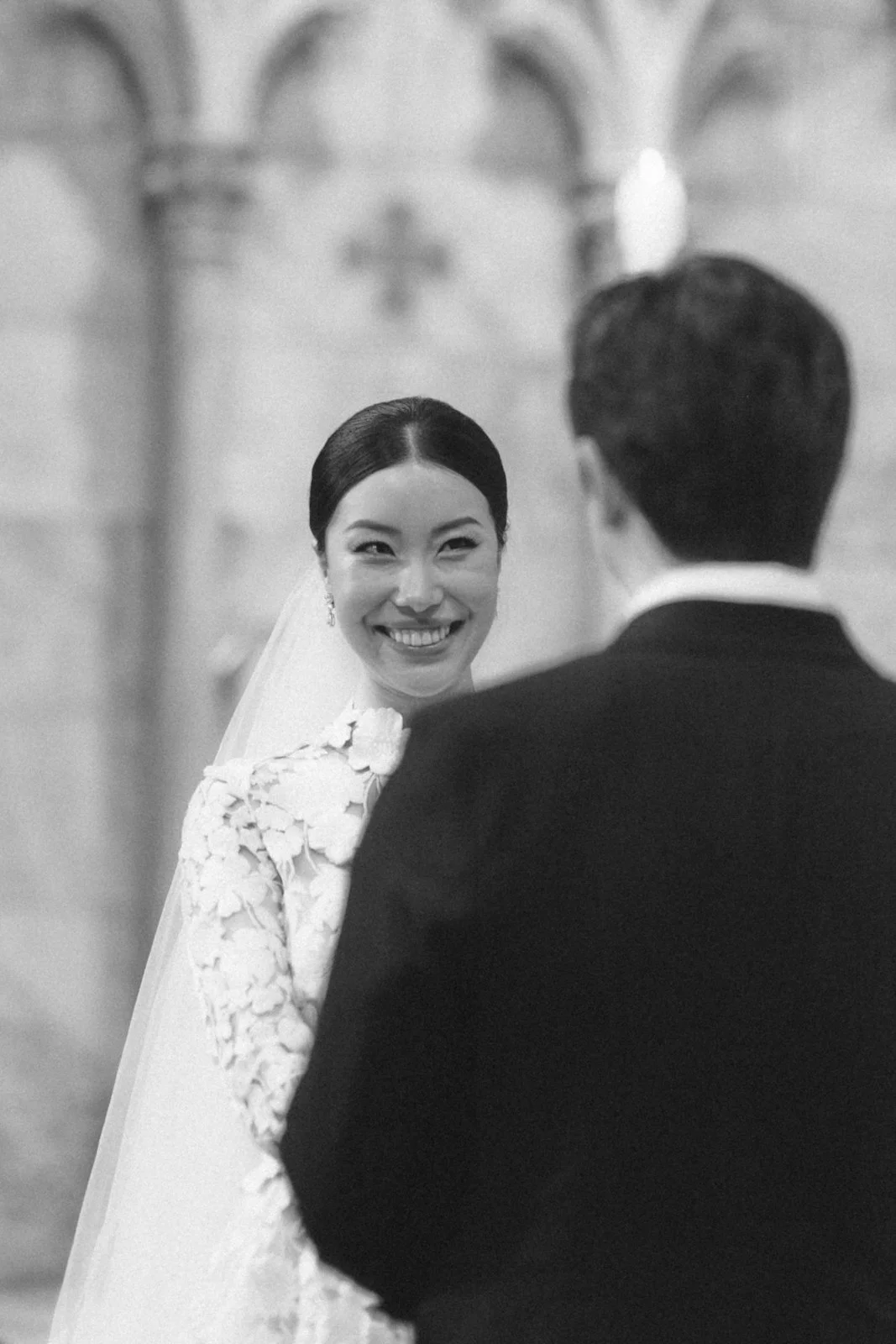Black and white photo of a bride smiling at a groom during their wedding ceremony in a church.