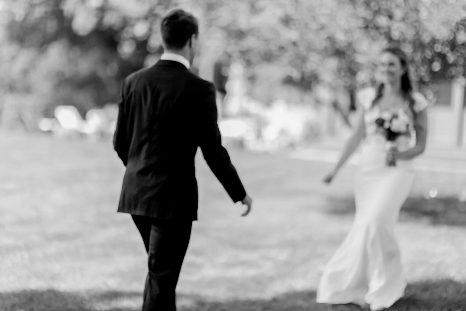 A black and white photograph of a man in a tuxedo walking towards a woman in a white evening gown holding a bouquet of flowers in an outdoor setting with trees in the background.