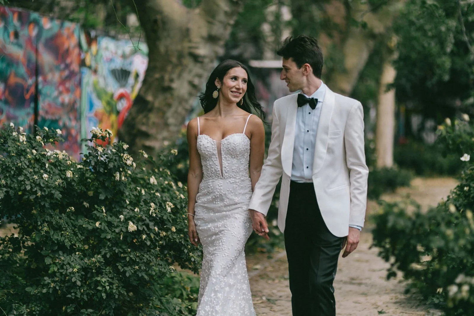A bride and groom walking hand in hand outdoors with colorful graffiti in the background.