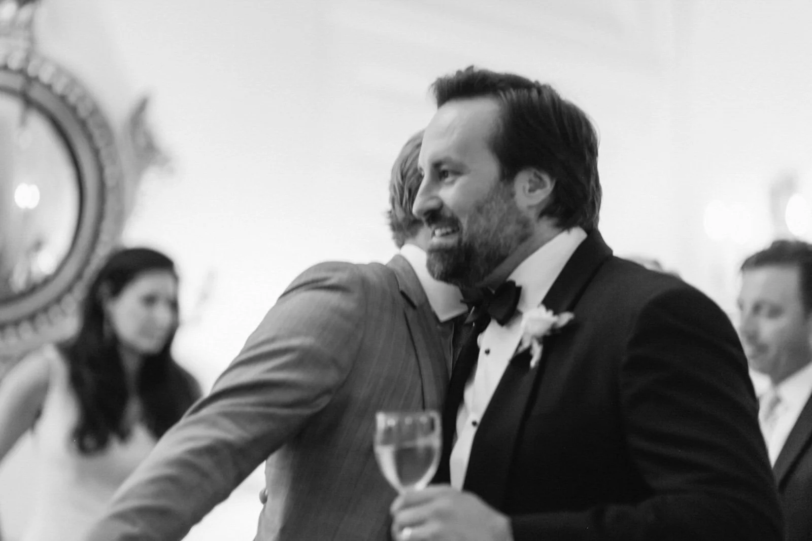 Black and white photo of a man with dark hair and a beard in a tuxedo with a bow tie, holding a glass of champagne at a formal event, smiling and embracing another man.