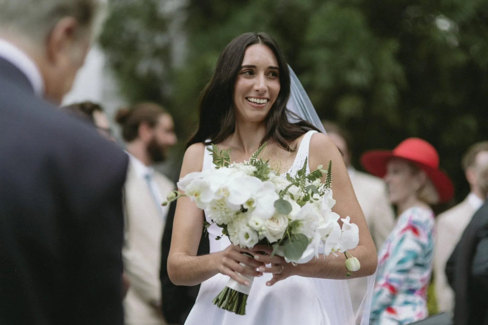 A bride in a white wedding dress holding a bouquet of white flowers and greenery, smiling at her wedding ceremony outdoors with guests in the background.