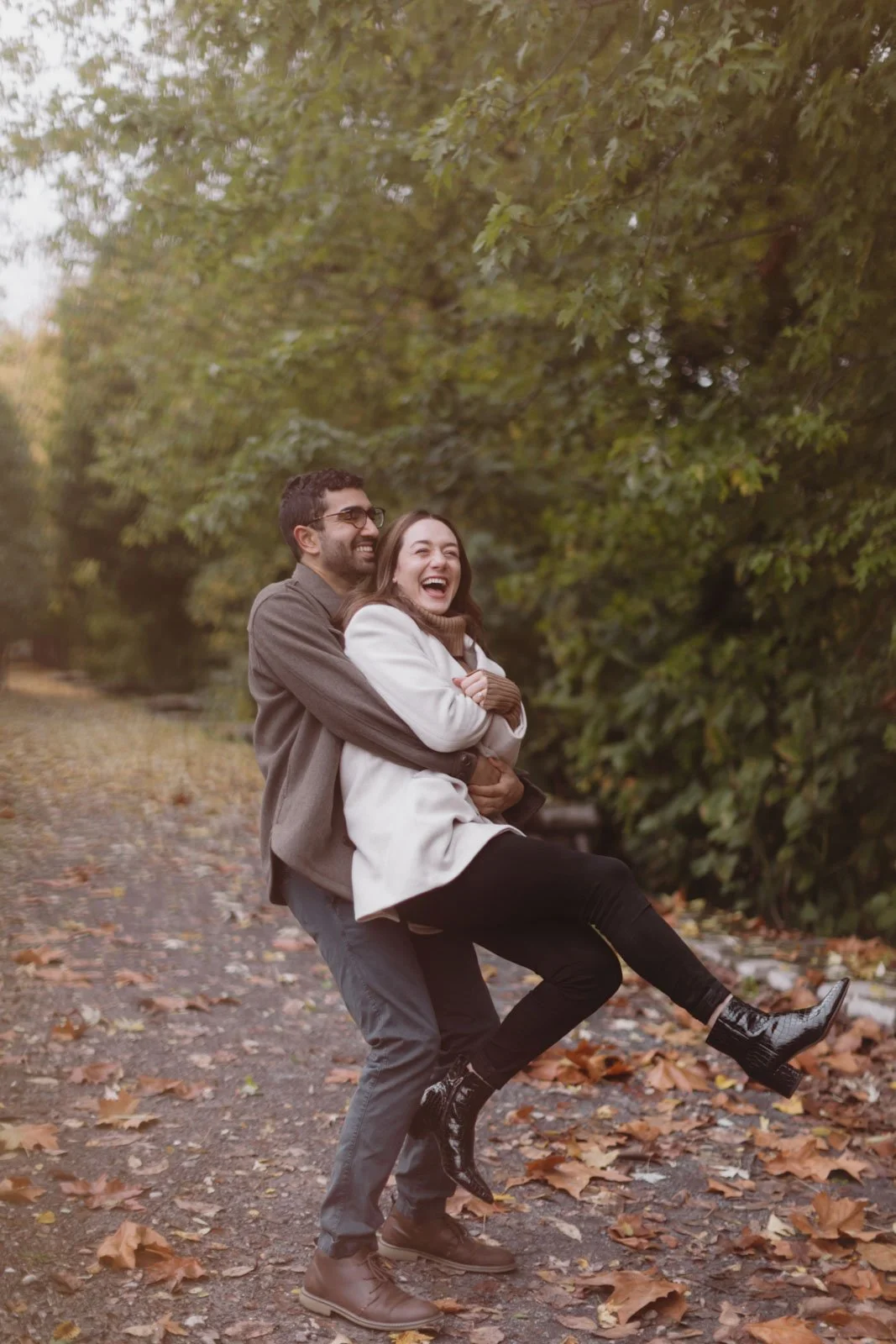 A man and woman are outdoors, laughing and hugging, with the woman being held in the man's arms. They are standing on a leaf-covered path surrounded by green trees and fall foliage.