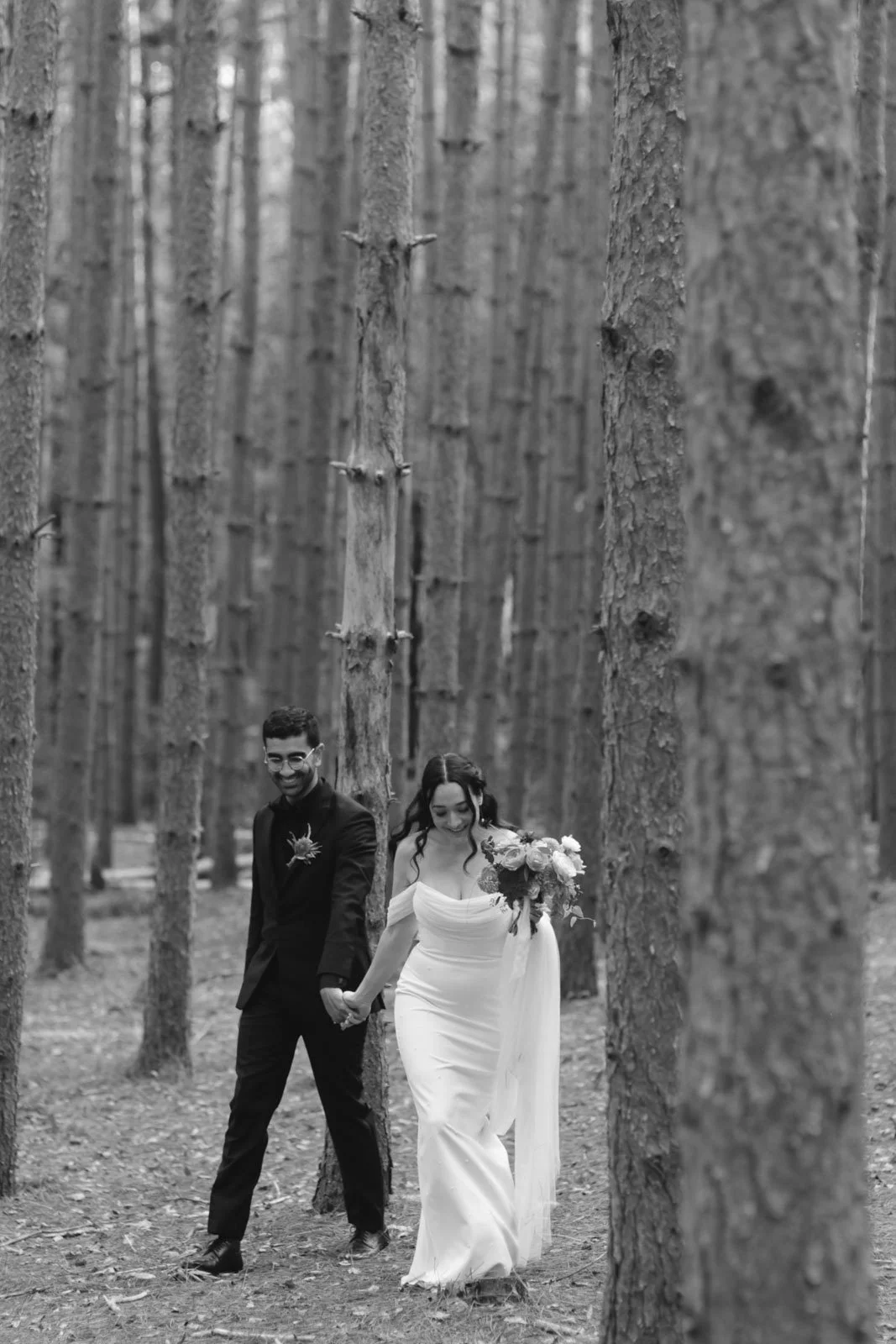 A couple holding hands and walking in a forest, with both smiling, the woman holding a bouquet of flowers.