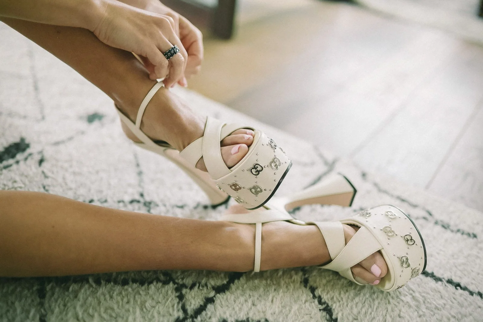 A woman putting on cream-colored high-heeled sandals decorated with metallic embellishments while sitting on a textured rug.