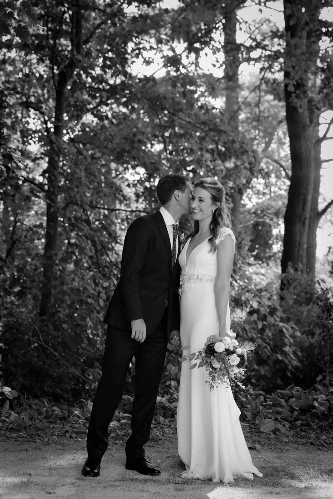 Black and white photo of a bride and groom in a wooded outdoor setting. The groom is whispering in the bride's ear, who is holding a bouquet of flowers and smiling.