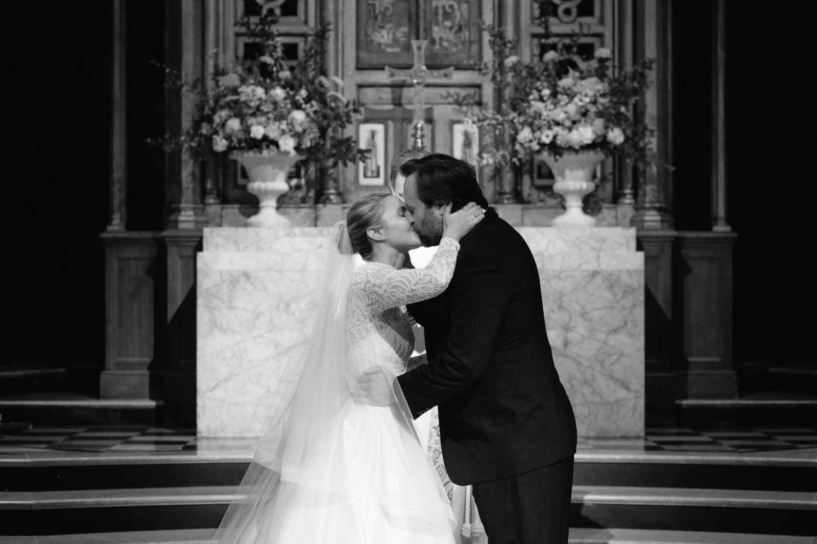 A bride and groom sharing a kiss during their wedding ceremony inside a church with floral arrangements and an altar in the background.