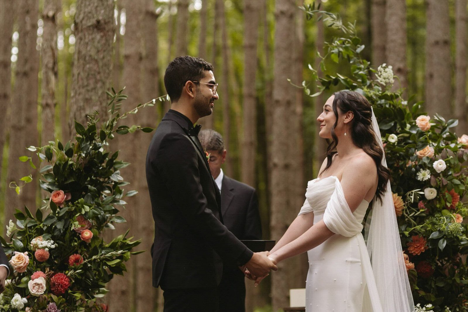 A couple getting married in a forest, holding hands and smiling at each other, with flowers and greenery surrounding them at Roxbury Barn and Estate.