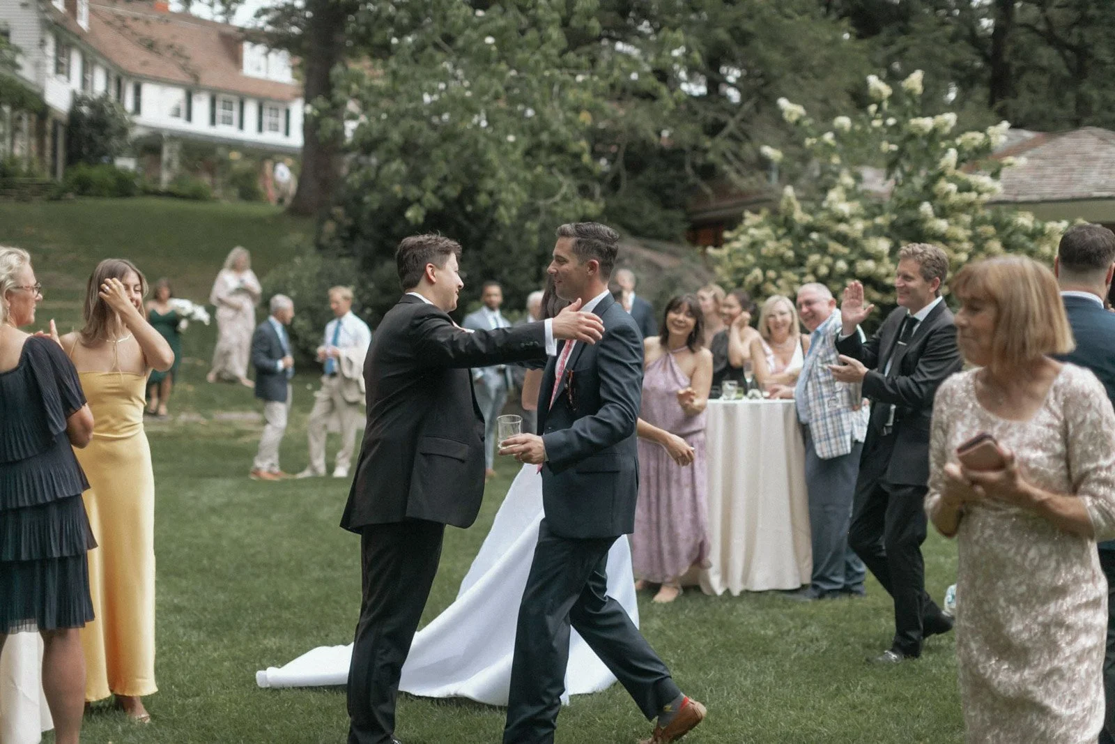 People dancing and socializing outdoors at a wedding reception, with a bride in a white gown and guests in formal attire enjoying the celebration under trees and on a grassy lawn.