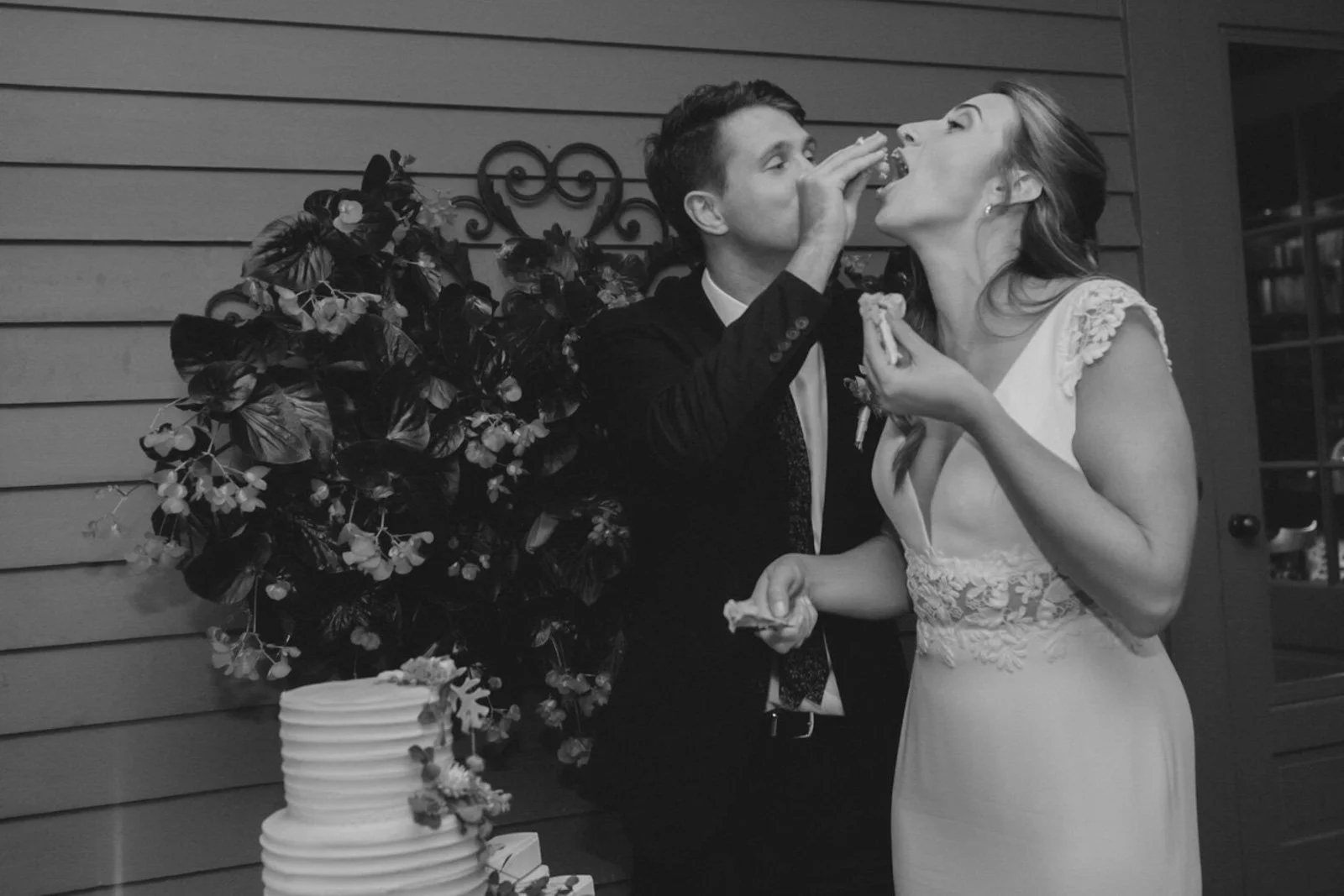 Black and white photo of a wedding couple feeding each other cake at a reception. The bride wears a lace dress, and the groom is in a suit. There is a wedding cake and decorative flowers in the background.