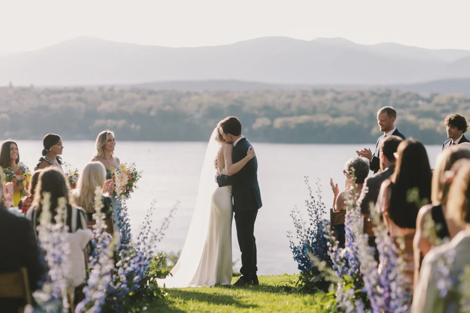 A bride and groom kissing during an outdoor wedding ceremony by a lake with mountains in the background, surrounded by seated and standing guests.