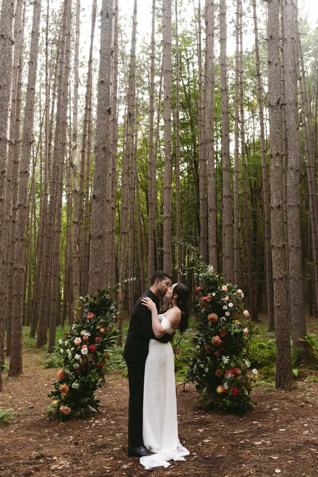 A couple in wedding attire, kissing in a wooded forest clearing with floral arrangements on either side.