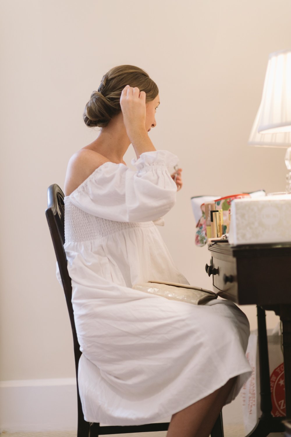 A woman sitting at a vanity table, applying makeup, in a white off-the-shoulder dress with puffed sleeves.