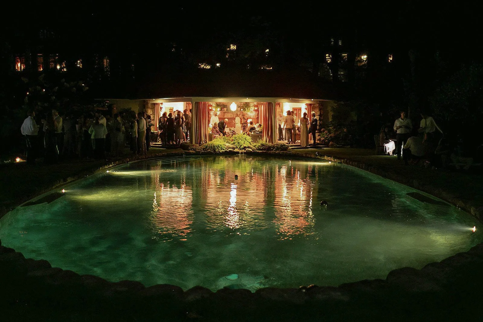 Nighttime scene of a gathering of people inside a house with large windows, with a reflective pool of water in the foreground and warm interior lighting.