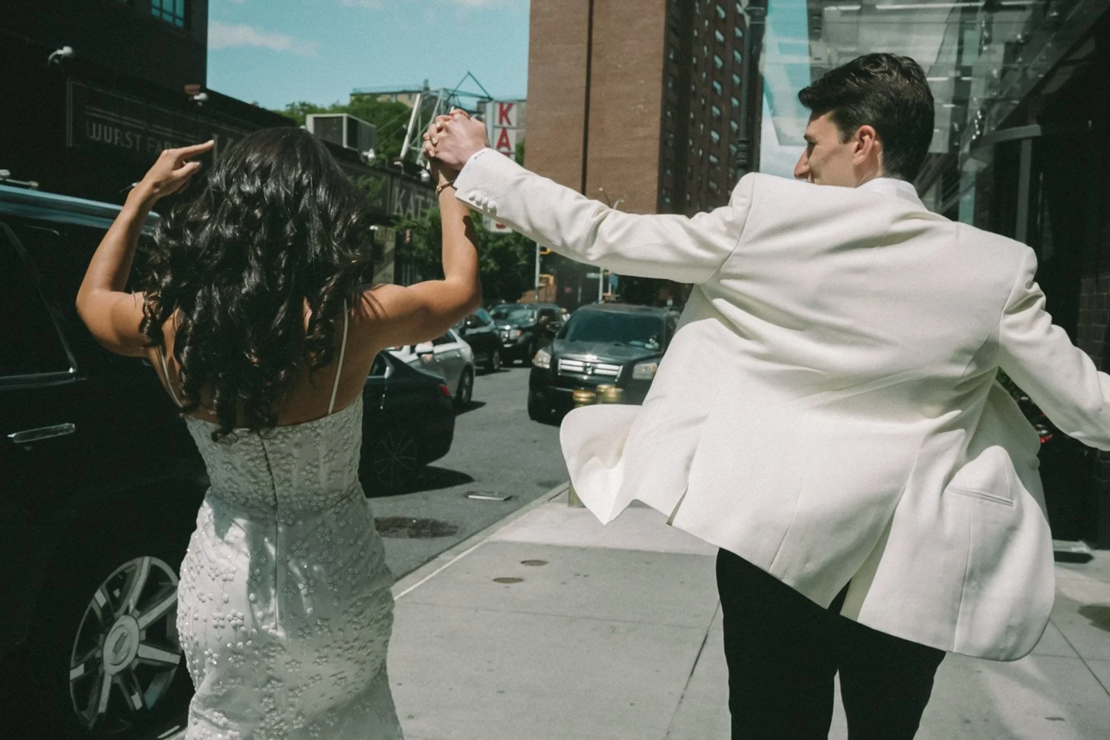 A couple dancing on a city sidewalk, the woman wearing a white dress with floral embellishments and the man in a white blazer and black pants, with cars and buildings in the background.