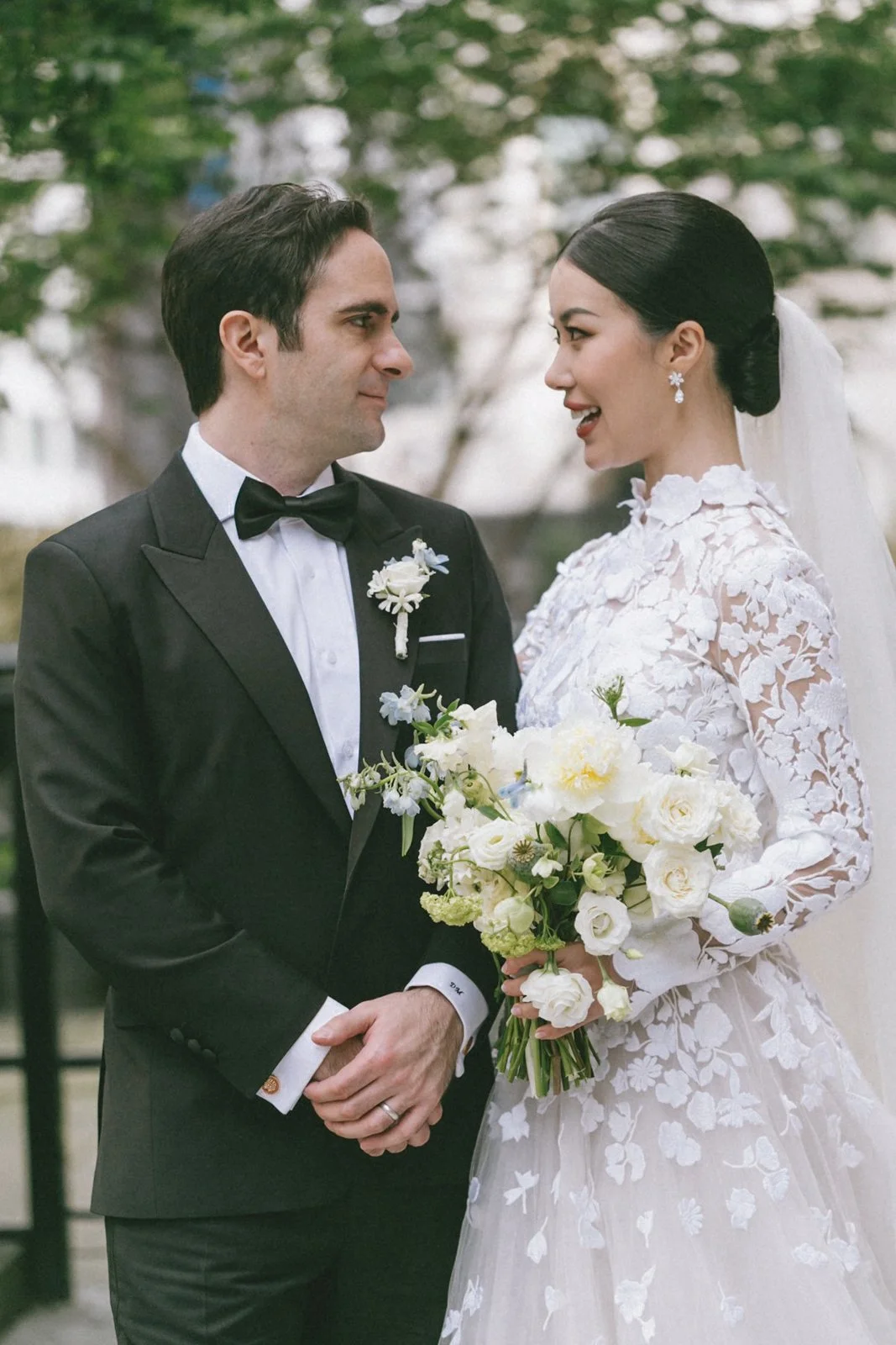 A bride and groom facing each other on their wedding day outdoors, with the bride holding a bouquet of white flowers and wearing a lace wedding dress, and the groom dressed in a black tuxedo with a bow tie, sharing a moment of connection.