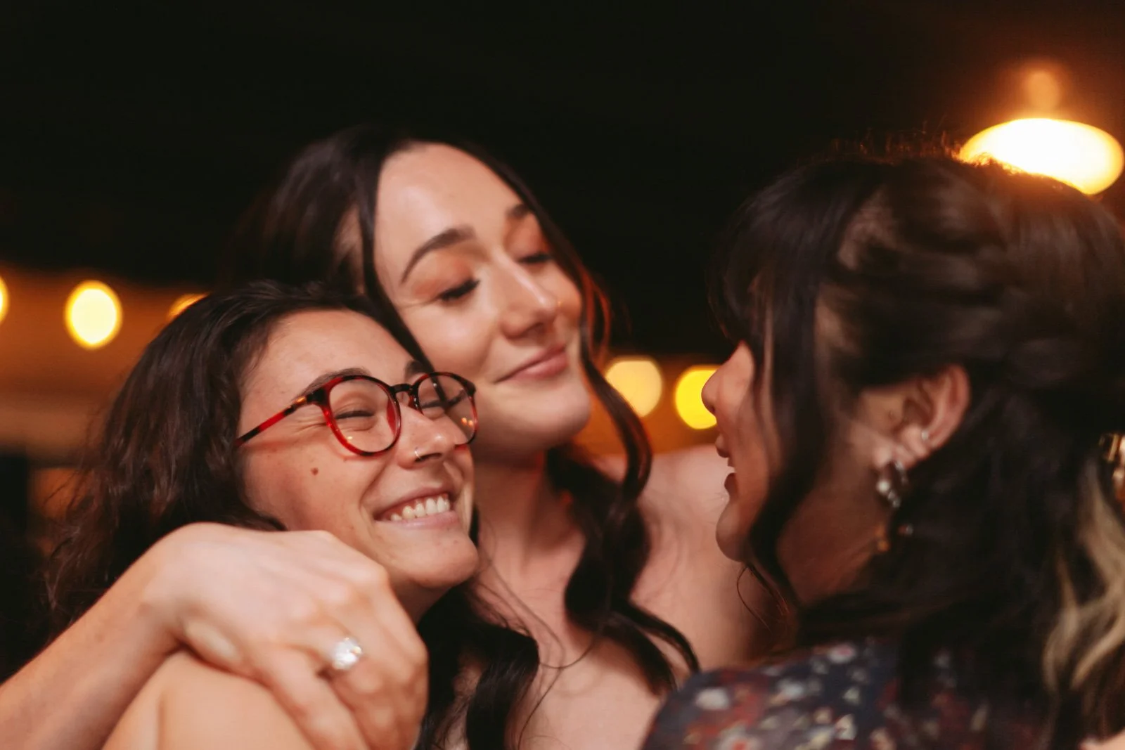Three women hugging and smiling at a social gathering with warm lighting in the background.