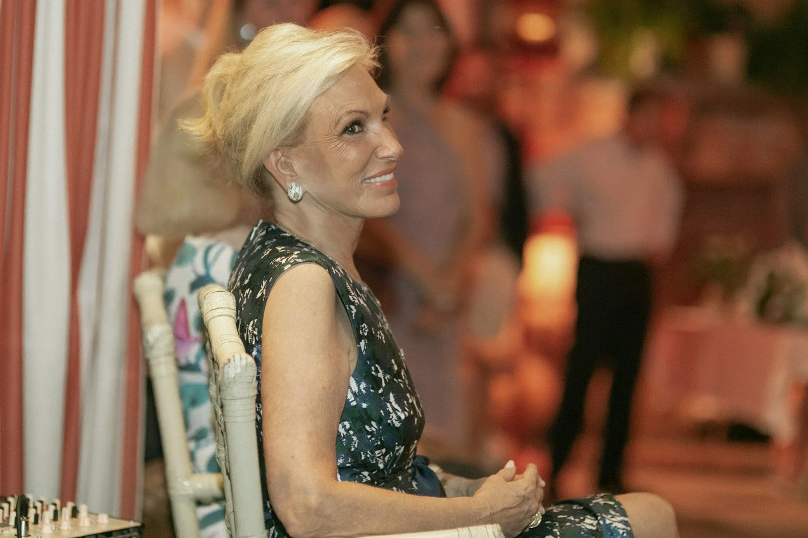 A woman with short blonde hair, wearing a floral sleeveless dress and earrings, sitting on a white chair in a warmly lit indoor setting with blurred people in the background.