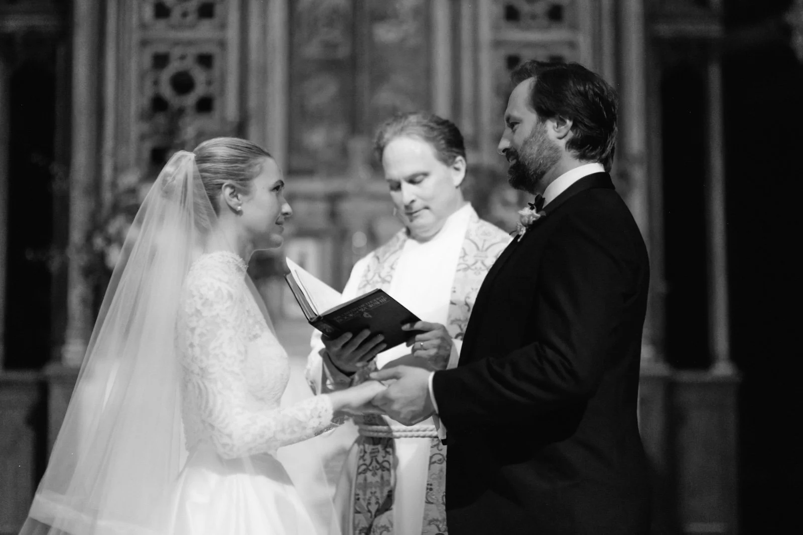 A black and white photo of a wedding ceremony inside a church. The bride and groom are holding hands, facing each other, with a priest or officiant reading from a book. The bride wears a lace wedding gown and veil, and the groom wears a tuxedo. The b