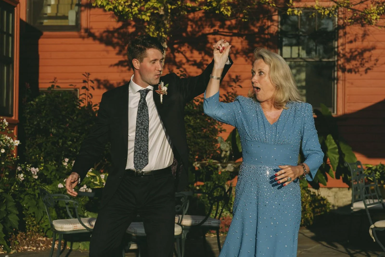 A man in a suit and a woman in a blue dress are dancing together outdoors during a celebration, with the man holding the woman's hand up while she looks surprised.